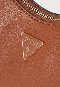Brun strukturerad läderhandväska med en triangulär metall-logobadge med texten "GUESS U.S.A." och en frågetecken-design. Guld dragkedjedetalj.