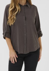 Chemise boutonnée gris foncé avec col et manches retroussables, en tissu légèrement texturé et ourlet arrondi.