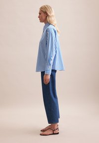 Camicia a righe blu e bianche a maniche lunghe con colletto alto, abbinata a pantaloni ampi in denim scuro e sandali neri flat.