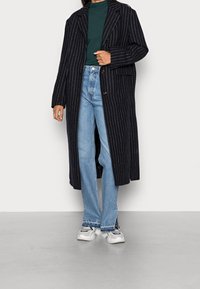 Femme portant un long manteau noir à fines rayures, un haut vert, un jean bleu clair fendu à l'avant, et des baskets blanches sur un fond clair uni.