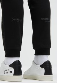 Schwarze Jogginghosen mit Logo-Prints, kombiniert mit weißen Sneakers, die eine schwarze Fersenpartie haben, und knöchellangen weißen Socken.