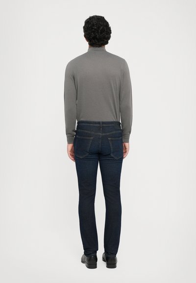 Man standing facing away wearing a gray long-sleeve shirt, dark blue jeans, and black shoes against a plain white background.