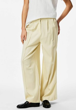 Personne portant un pantalon ample de couleur crème, un haut blanc et des mocassins noirs, debout devant un fond uni.