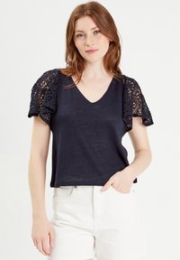 Marineblauwe top met korte mouwen, een V-hals en kantdetails op de mouwen; de stof is lichtgewicht met een relaxte pasvorm, gecombineerd met witte shorts.