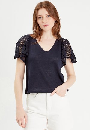 Haut à manches courtes bleu marine avec décolleté en V et détails en dentelle sur les manches ; tissu léger avec une coupe décontractée, associé à un short blanc.