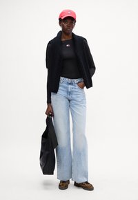 Gilet nero a maglia sopra una maglietta nera a maniche lunghe, jeans flare blu chiaro, scarpe con stampa leopardata e un cappellino rosso. Tiene una borsa a tracolla nera.