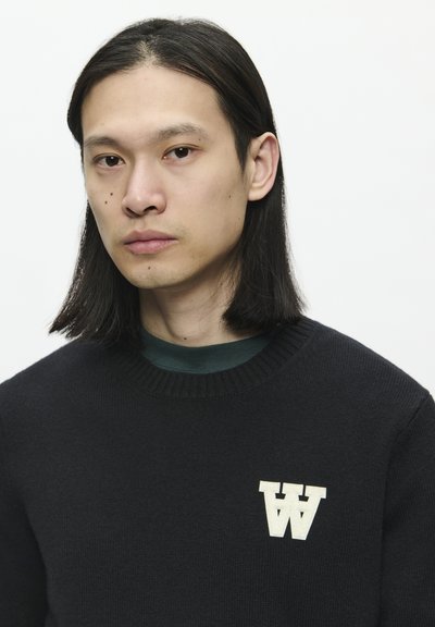 Suéter negro de punto con cuello acanalado, que presenta un logo blanco de "W" en el pecho izquierdo. Debajo, se puede ver una camisa de cuello redondo en verde oscuro.
