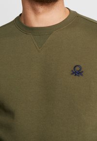 Sudadera verde oliva con cuello acanalado y acentos en los hombros. Presenta un pequeño logo bordado en azul marino en el pecho. Textura suave.