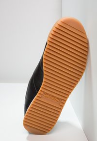 Semelle de chaussure Reebok noire avec une bande de roulement en caoutchouc orange, présentant des rainures horizontales et le logo Reebok au centre.