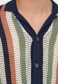 Acercamiento de una camisa de cuello de punto azul marino con franjas de crochet verticales en azul marino, terracota, blanco y verde salvia, abotonada en el frente.