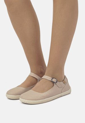 Pies que llevan zapatos estilo Mary Jane en color beige con hebillas y suelas de goma blanca sobre un fondo blanco liso.