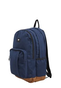 Marinblå tygryggsäck med två huvudfack med dragkedja, brun läderbotten, sidofickor och en logopatch på framsidan.