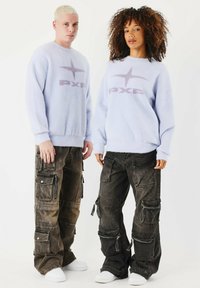 Pullover fuzzy blu chiaro con logo viola, abbinato a pantaloni cargo scuri con molteplici tasche e cerniere. Sneakers bianche completano il look.