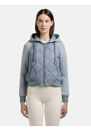 Kvinde iført lys blå hættequiltejacket med snore og beige leggings, stående med fronten mod kameraet mod en ensfarvet baggrund.