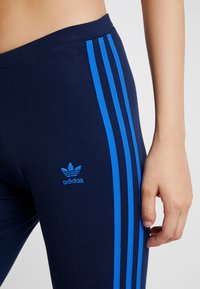 Marina blå leggings med tre djärva blå ränder längs sidan och en liten blå adidas-logotyp nära midjan, tillverkade av ett elastiskt tyg.