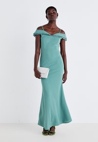 Rochie maxi teal cu umeri goi, cu detalii din dantelă, bust ajustat și fustă evazată. Modelul poartă o poșetă argintie și sandale verzi cu bretele.