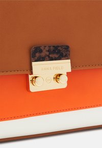 Brązowo-pomarańczowa torba z ekoskóry z złotym zatrzaskiem. Posiada teksturowany ciemny akcent i logo "ANNA FIELD". Płaski projekt z kontrastującymi kolorami.