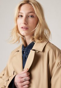 Impermeabile beige con un design classico, caratterizzato da una chiusura con bottoni e colletto. Sotto viene indossata una camicia di jeans blu. Pelle lentigginosa e capelli chiari.