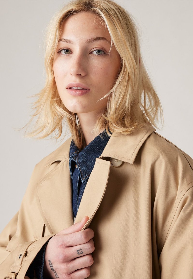 Beige Trenchcoat mit klassischem Design, featuring einer knopfbaren Front und einem Kragen. Darunter wird ein blauer Denim-Hemd getragen. Sommersprossige Haut und helles Haar.