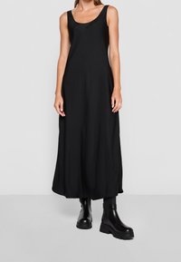 Vestido maxi negro sin mangas combinado con botas de tobillo de cuero negro grueso, mostrado en una persona de pie frente a un fondo liso.