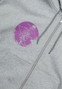 Felpa grigia con zip che presenta una grafica viola di uno scheletro all'interno di un design circolare. Il tessuto sembra morbido e testurizzato.