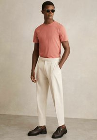 Tricou roz corali cu mâneci scurte, pantaloni crem ajustați și pantofi maro închis. Detalii de accent includ o textură netedă și o croială structurată.