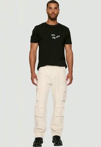 Homem de pé vestindo uma t-shirt gráfica preta, calças cargo bege com múltipos bolsos e sapatos pretos, contra um fundo neutro.