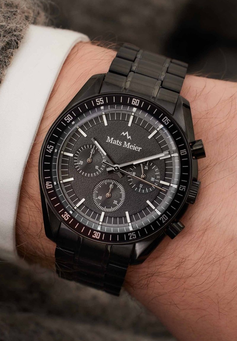 Zwarte chronograafhorloge met een gestructureerde donkere wijzerplaat, zilveren accenten en een metalen band. Heeft drie subwijzerplaten en een tachymetrische schaal.