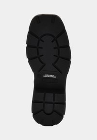 Suola di scarpa in gomma nera con superficie texture, modellata con scanalature e motivi profondi per una migliore trazione. Presenta un'etichetta con informazioni sul prodotto.