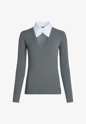 Maglione grigio a coste con maniche lunghe e scollo a V profondo, indossato sopra una camicia bianca con colletto appuntito, con piccolo dettaglio ricamato vicino all’orlo.