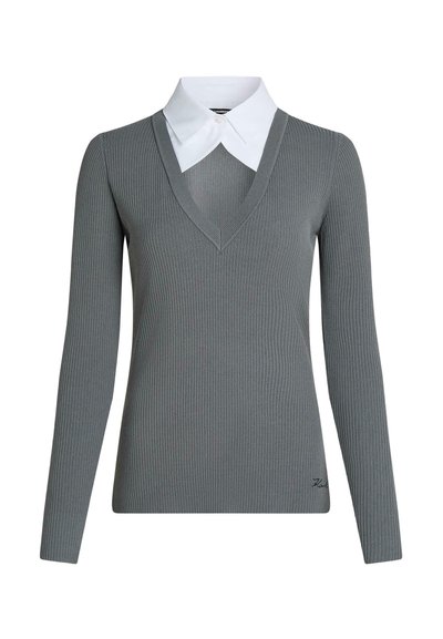 Pull gris côtelé à manches longues avec un profond col en V, superposé à une chemise blanche à col pointu, avec un petit détail brodé près de l'ourlet.