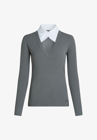 Pull gris côtelé à manches longues avec un profond col en V, superposé à une chemise blanche à col pointu, avec un petit détail brodé près de l'ourlet.