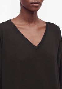 Gros plan d'une personne portant un pull en maille noire à col en V, montrant le cou, la clavicule et le haut de la poitrine.