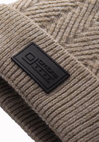 Gorra de punto beige con textura acanalada, que presenta un parche rectangular de cuero negro etiquetado "ombre 1985" en el puño.