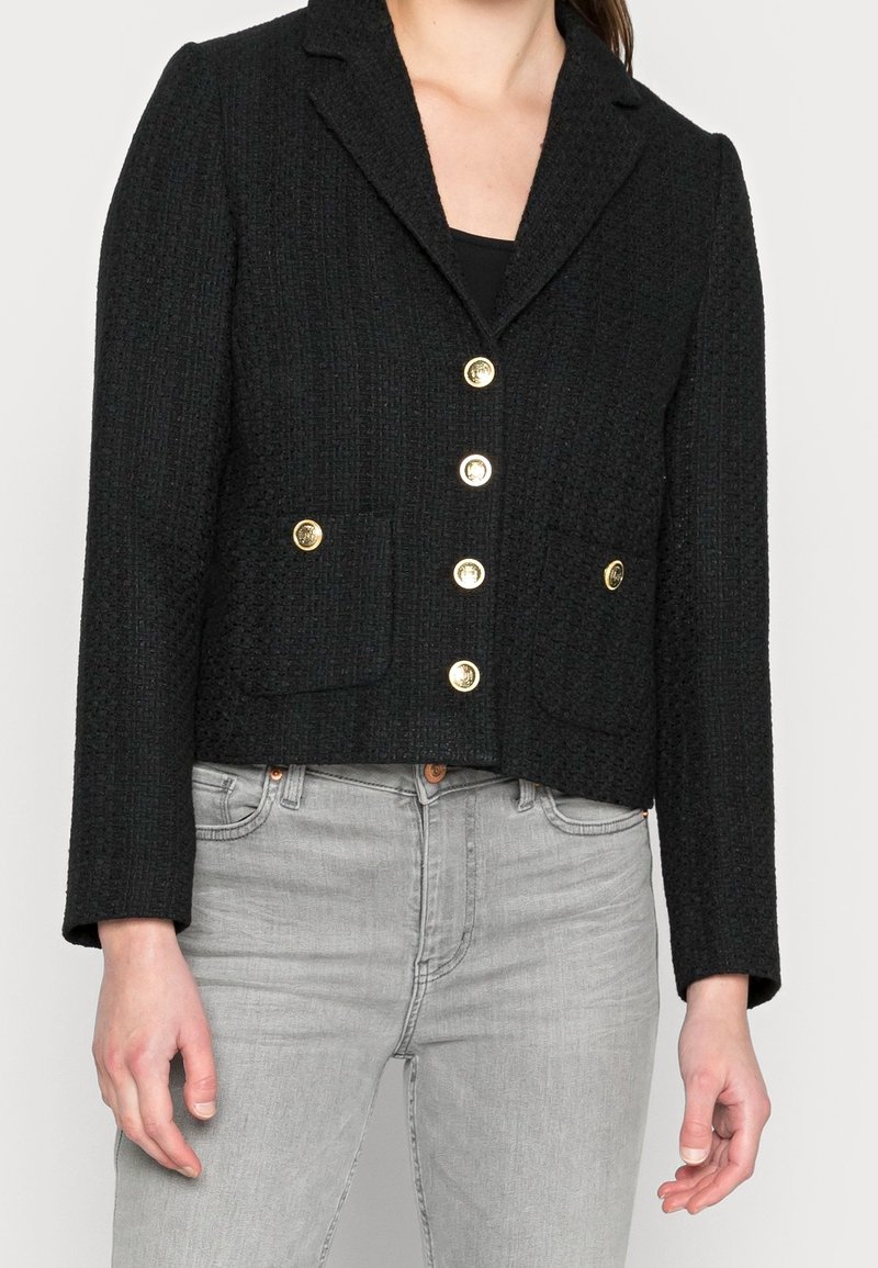 Femme portant un blazer noir texturé avec des boutons dorés sur un top noir et un jean gris clair.