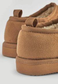 Zapatillas deslizantes de color marrón con textura y un interior suave, que cuentan con un perfil bajo, suela plana de goma y pequeñas cintas de tela en el talón.