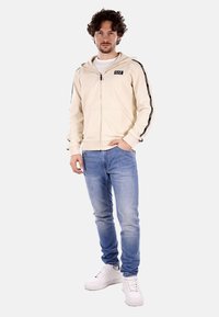 Felpa beige con zip, strisce laterali nere e logo, abbinata a jeans azzurri e sneakers bianche. Abbigliamento sportivo casual.