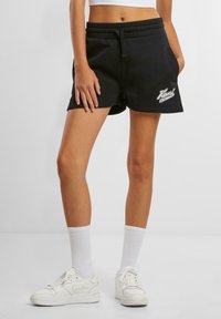 Schwarze Baumwollshorts mit einem elastischen Bund und Kordelzug, versehen mit einem weißen Grafiklogo auf der Vorderseite. Kombiniert mit weißen Turnschuhen und Socken.