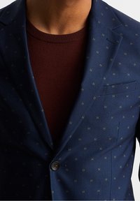 Navy blazer with subtle gold geometric pattern, single button closure, and notch lapels, layered over a maroon sweater.