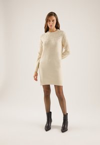 Beige knitted sweater dress with a round neck, embellished with small pearls on the front and long sleeves. Worn with black ankle boots.