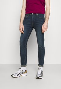 Mörkblå slim-fit jeans med femfickdesign, rullade fållar, kombinerat med silver- och svarta sportskor samt en magentafärgad t-shirt.