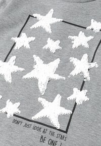 Grauer Stoff mit einem Pailletten-Stern-Design in Weiß. Besitzt mehrere Sternformen, die von einem schwarzen Rechteck umschlossen sind, und enthält Text am unteren Rand.