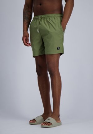 Man draagt olijfgroene zwembroek en bijpassende slidesandalen, staat met één hand in de zak, neutrale achtergrond.