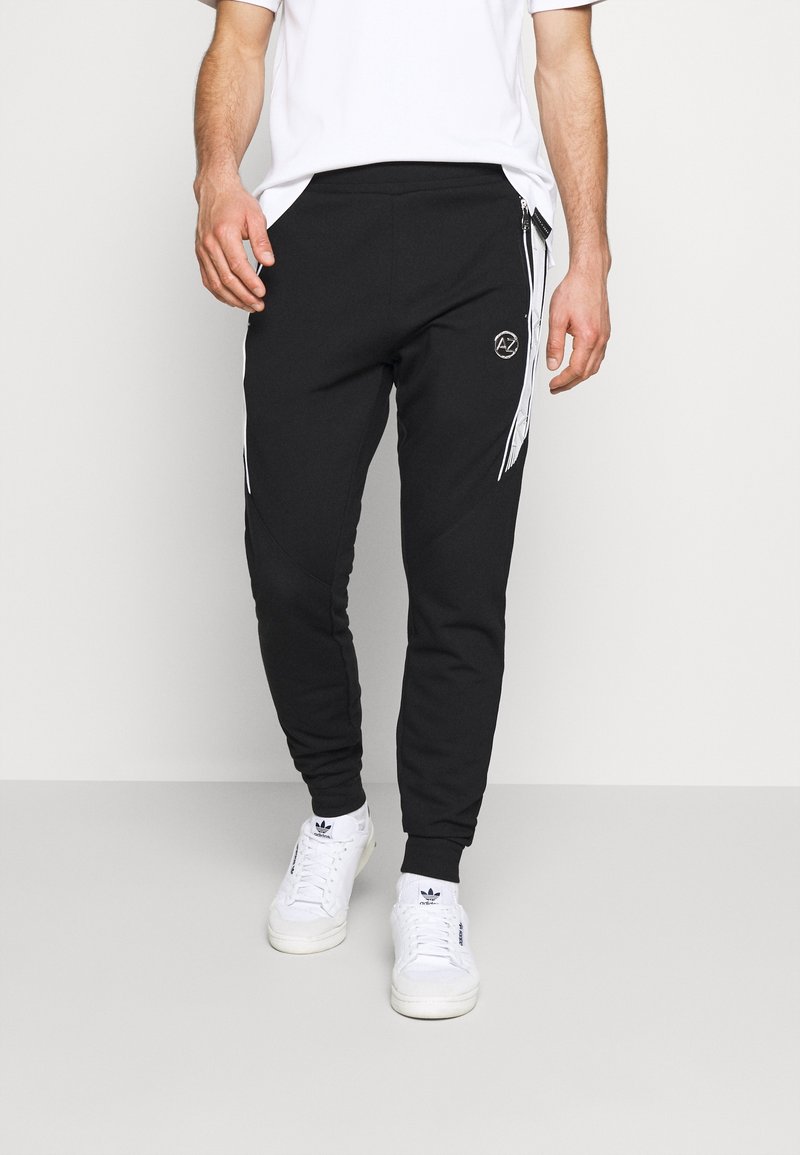 Černé joggingové kalhoty vyrobené z měkkého materiálu. Mají bílé boční pruhy, vyšitý logo a zúžený střih nohavic s elastickými manžetami.