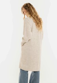 Vrouw met lang krullend haar draagt een beige gebreide cardigan met zakken, gecombineerd met blauwe jeans, terwijl ze tegen een witte achtergrond staat.