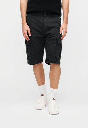 Mand iført sorte cargobukser, hvide sokker med logo og hvide sneakers, står foran en ensfarvet lys baggrund.