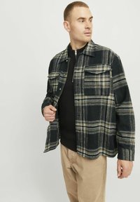 Cămașă din flanel cu carouri în nuanțe închise de albastru și verde, cu guler, două buzunare pe piept și închidere cu nasturi, purtată peste un pulover negru tricotat.