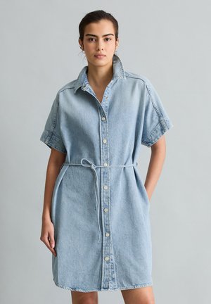 Jonge vrouw draagt een lichtblauwe denimjurk met korte mouwen, knopen aan de voorkant en een dunne riem gestrikt in de taille, staand tegen een effen grijze achtergrond.