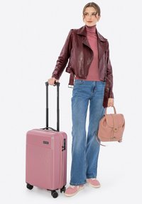 Roze harde koffer met een textuurovervlak, zwarte telescoop handgreep en draaiwielen. Gecombineerd met een lichtroze rugzak en een casual outfit.