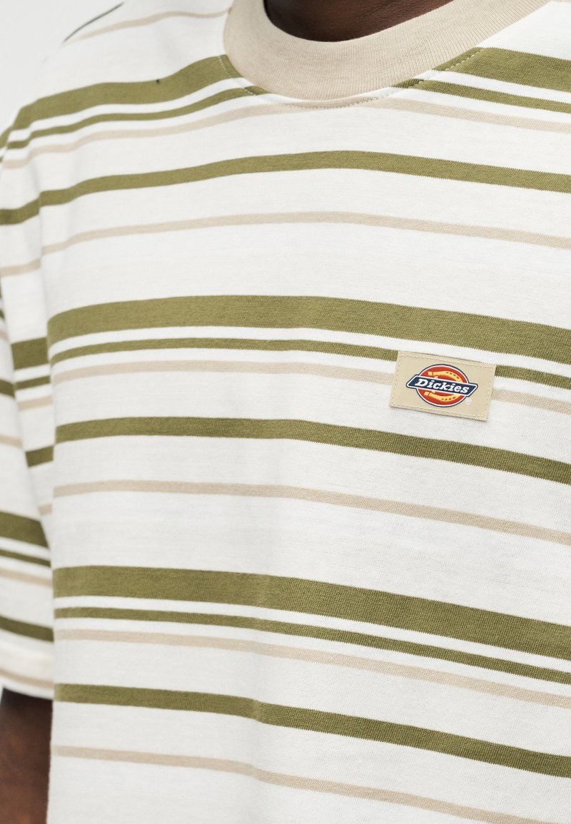 Nahaufnahme einer Person, die ein weißes T-Shirt mit beigen und olivgrünen horizontalen Streifen und einem kleinen Dickies-Logo-Aufnäher auf der Brust trägt.